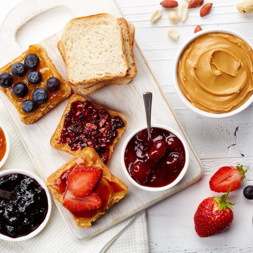 Sandwiches with peanut butter, jam and fresh fruits on white wooden background from top view